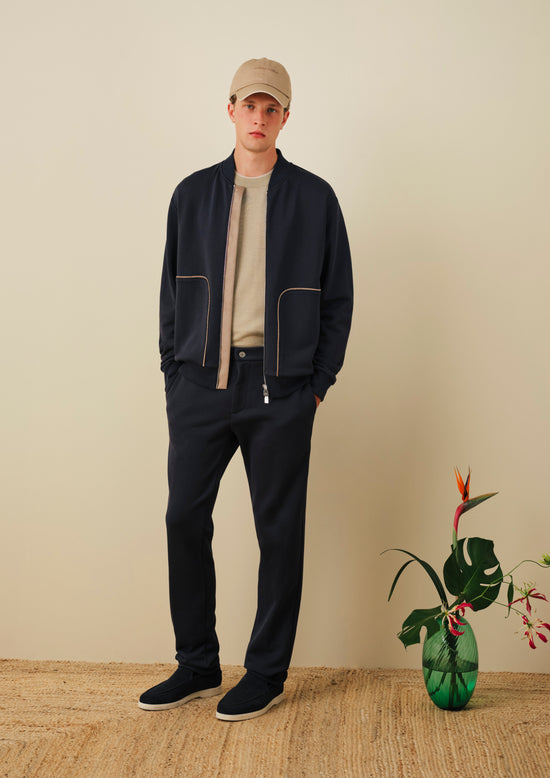 Man wearing navy blue bomber jacket with contrast piping, beige t-shirt, navy trousers, tan baseball cap, and black sneakers, standing against beige wall with decorative vase