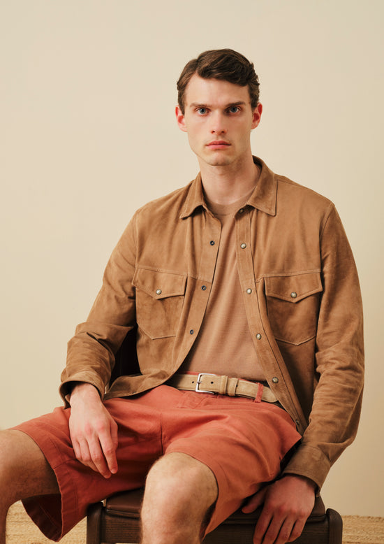 Man sitting wearingsuede camel brown overshirt with chest pockets, terracotta shorts, and matching belt against beige background