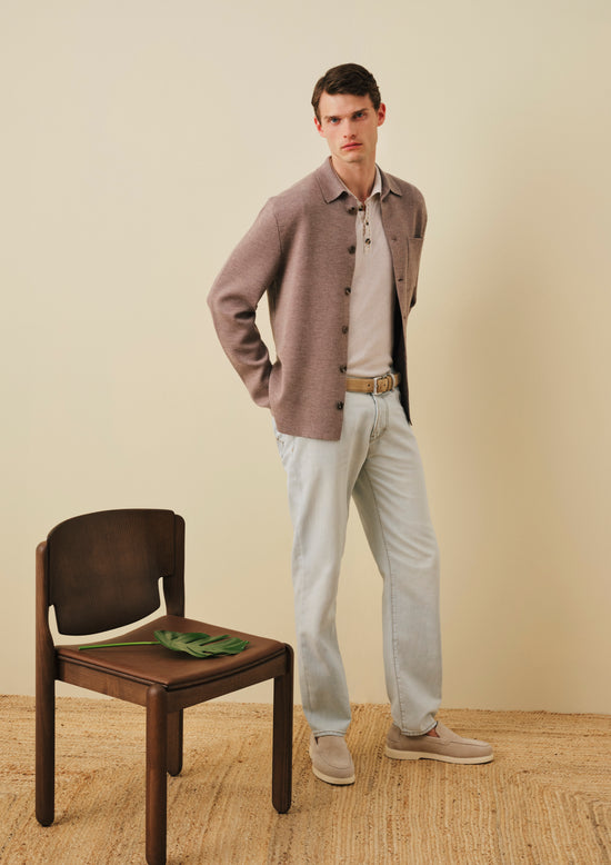 Man wearing dusty pink wool overshirt over beige polo, light blue jeans and brown leather belt with beige suede loafers, standing next to wooden chair against neutral background