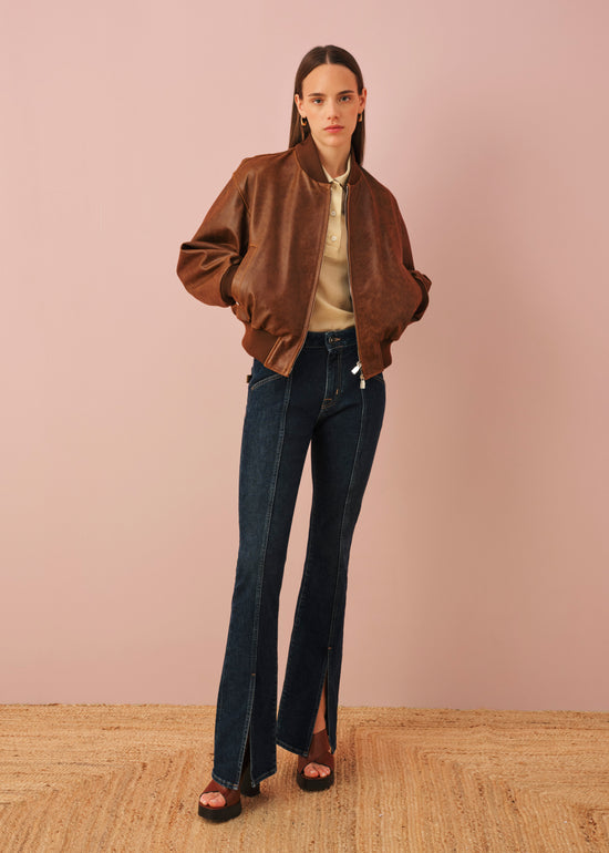 Woman wearing brown bomber jacket over cream hoodie with dark blue flared jeans and black sandals against pink background