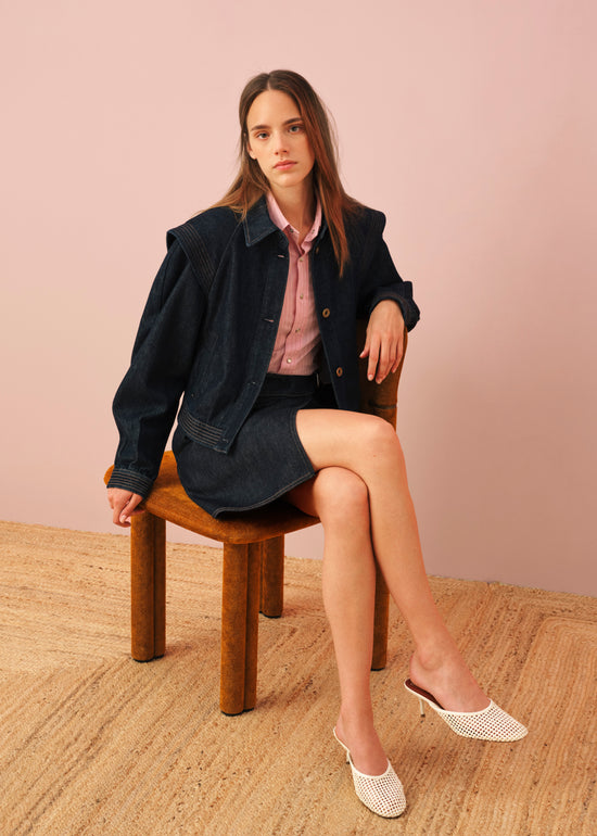Woman sitting on wooden stool wearing navy denim jacket and skirts, pink shirt, and white woven leather mules against pink background