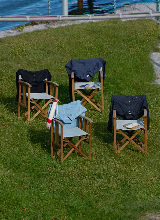 Four wooden folding chairs arranged on a grass lawn by a waterfront, each displaying a different shirt in shades of navy, blue and light blue