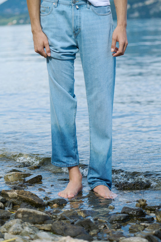 Close-up of a person standing barefoot in shallow water on a rocky shore, wearing light wash straight fit jeans.