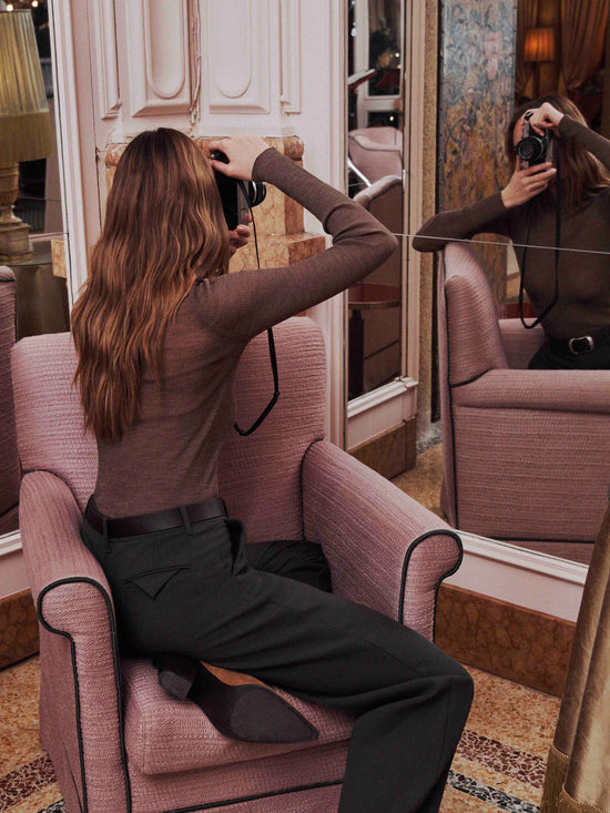 Woman sitting on pink armchair in elegant boutique taking mirror selfie wearing black tailored trousers and brown knit top