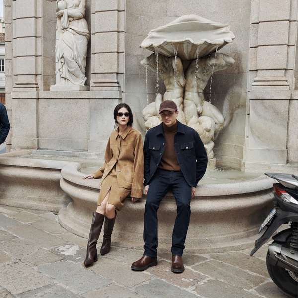 Two models in European city square by baroque fountain: man in denim jacket and olive trousers, woman in camel suit with brown boots