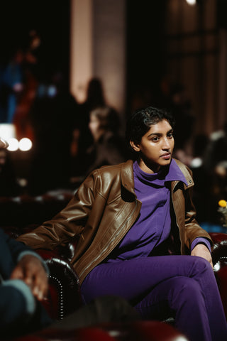 Guest seated on a brown Chesterfield armchair in the evening lounge of a five-star hotel: a woman wearing a deep violet outfit and a glossy brown leather bomber jacket with a collar