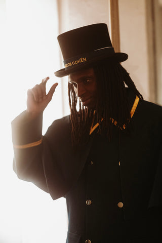 Close up portrait of a doorman in full five star hotel uniform: wearing a top hat with a gold band and the embroidered name “Jacob Cohën” in gold, a navy blue double breasted jacket with gold epaulettes and braid, and shiny gold buttons