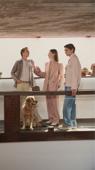 Group of three models wearing neutral-toned luxury clothing including blazers, shirts and trousers in modern gallery space with wooden ceiling and golden retriever