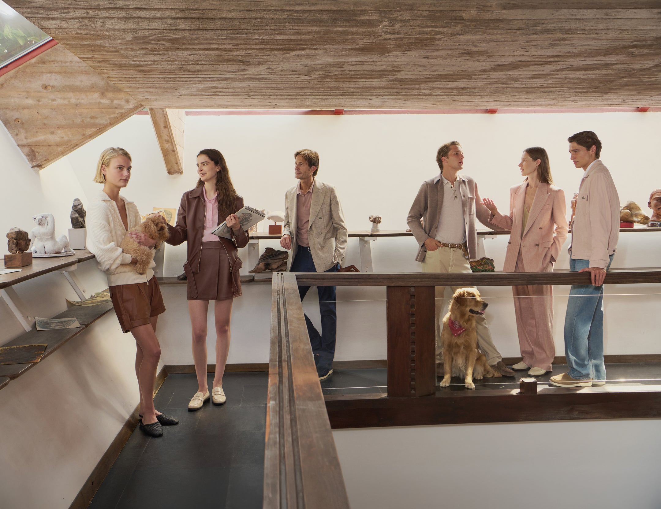 Group of six models wearing neutral-toned luxury clothing including blazers, shirts, shorts, and trousers in modern gallery space with wooden ceiling and golden retriever