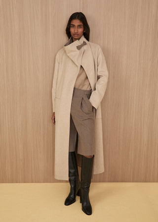 Model wearing a long asymmetric ivory double cashmere-silk wrap coat with a leather buckle closure at the high collar, a taupe top, a matching wrap skirt-shorts, and high black leather boots