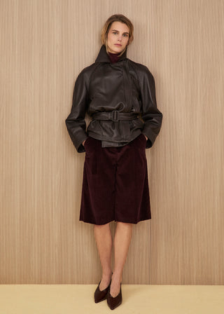 Dark brown soft leather jacket with a high wrap collar and a matching buckled waist belt, wide knee-length shorts in dark burgundy corduroy, and dark pointed heeled shoes