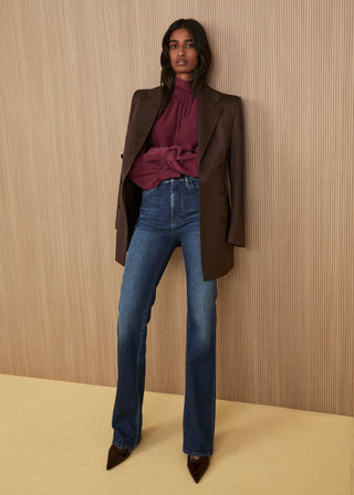 Model standing wearing a dark brown double-breasted blazer over her shoulders, a plum high-neck blouse with draped sleeves, dark blue wash high-waisted jeans, and pointed dark brown shoes