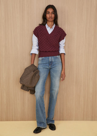 Model wearing a dark plum sleeveless textured v-neck knit vest over a blue and white striped shirt, light-wash regular flare jeans, black flat shoes, and holding a brown micro-check blazer
