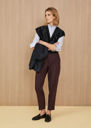 Black textured v-neck knit vest worn over a light blue and white striped shirt, dark jacket held in hand, dark burgundy tailored cuffed trousers, and black leather loafers