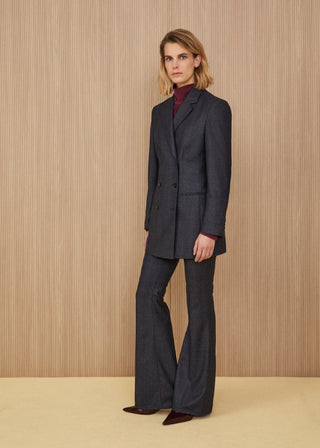 Model wearing a dark charcoal grey tailored double-breasted pantsuit featuring a structured blazer and matching flared trousers, paired with a plum turtleneck top and dark pointed shoes