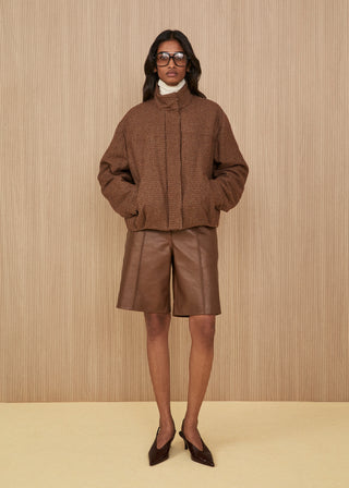 Model wearing glasses features a padded high-neck puffer jacket in a brown and beige micro-check pattern, wide knee-length brown leather shorts, and dark brown pointed slingback heels