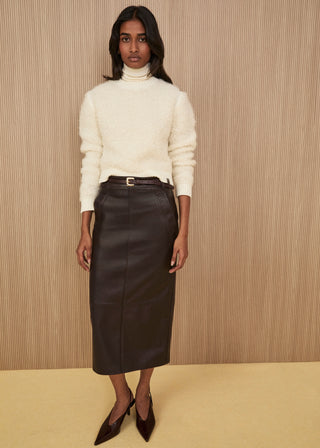 Model standing wearing a soft cream bouclé knit sweater over an ivory turtleneck, a dark brown soft leather pencil midi skirt with a slim belt, and dark brown pointed heels
