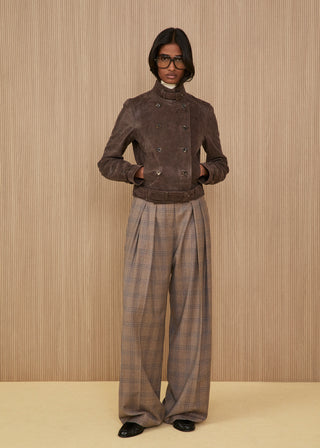 Model wearing glasses features a short double-breasted muddy brown suede jacket with a high structured collar and a belt, paired with fluid extra wide-leg trousers in a brown plaid pattern and black flat shoes