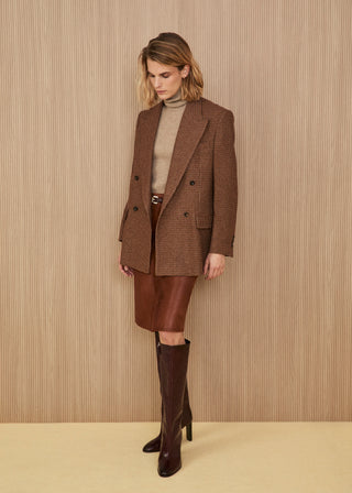 Model wearing a long double-breasted blazer in a brown and beige micro-check pattern, a taupe turtleneck sweater, a brown leather midi skirt with a belt, and high dark burgundy leather heeled boots