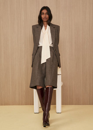 Model leaning on a design chair wearing a brown micro-check suit with a blazer and wide knee-length shorts, paired with an ivory silk blouse featuring a tie-neck and glossy high burgundy boots