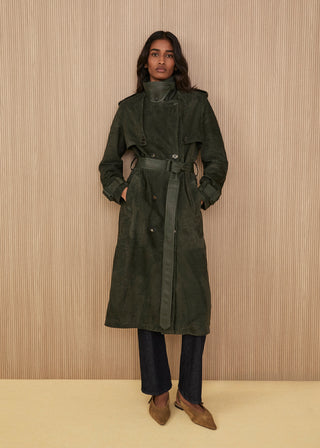 Model standing wearing a long double-breasted dark forest green suede trench coat with a matching belt, a structured high collar, dark indigo denim trousers, and pointed light brown suede shoes