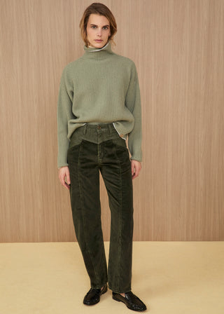 Model standing wearing a soft ribbed turtleneck sweater in sage green, dark forest green high-waisted corduroy trousers featuring a geometric yoke detail on the front, and glossy black leather loafers