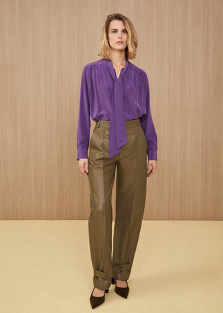 Model wearing a fluid silk blouse in electric purple with a lavallière tie-neck, high-waisted soft olive green leather trousers with ankle straps, and dark brown pointed heels