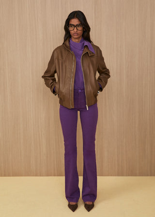Model wearing glasses features a zippered bomber jacket in soft brown leather, an electric purple high-neck blouse, and matching purple regular-fit flared jeans, finished with dark pointed heels