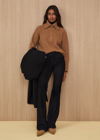 Model wearing a camel-colored ribbed sweater with a half-zip and high collar, flared black corduroy trousers, brown suede shoes, and holding a black coat