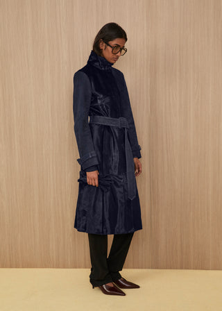 Model in profile wearing a long double-breasted belted coat in glossy dark indigo blue calf hair, featuring a high structured collar, paired with black straight-leg trousers and dark pointed heels