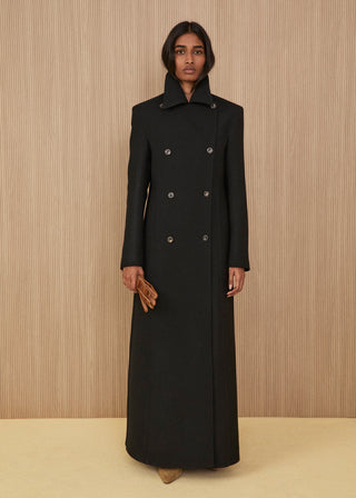Model standing wearing an ankle-length structured double-breasted black wool coat with defined shoulders and a closed high collar, holding camel-colored leather gloves