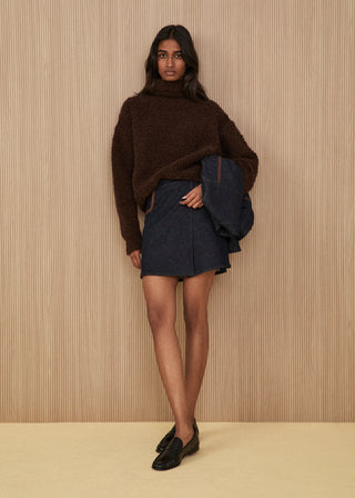 Dark brown bouclé knit high-neck sweater, dark indigo denim wrap miniskirt with brown leather piping, matching denim jacket held in hand, and black leather loafers
