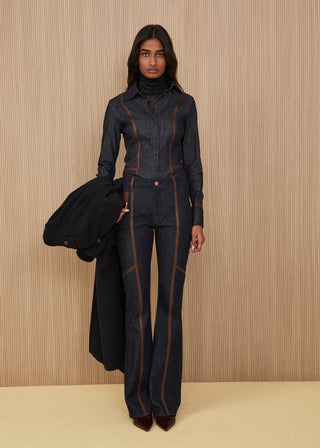 Dark indigo denim co-ord set featuring a structured denim shirt and matching regular jeans, both detailed with brown leather piping, a black coat held in hand, and pointed shoes