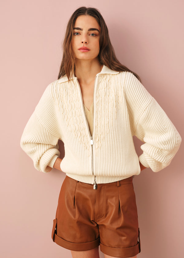 Model wearing a cream chunky ribbed-knit zip-up cardigan with cable-knit detail on the chest, paired with brown leather tailored shorts