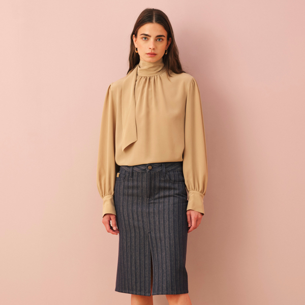 Model wearing a beige pussy-bow blouse with gathered neckline, puff sleeves and buttoned cuffs, styled with a dark navy blue striped denim midi pencil skirt with a front slit, against a soft pink background
