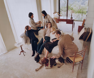 Group of six people in tailored neutral-toned knitwear and denim, relaxing together on a cream sofa in a bright living room, with a small poodle and a German Shorthaired Pointer dog, large windows overlooking greenery in the background