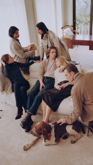 Group of six people in tailored neutral-toned knitwear and denim, relaxing together on a cream sofa in a bright living room, with a small poodle and a German Shorthaired Pointer dog, large windows overlooking greenery in the background