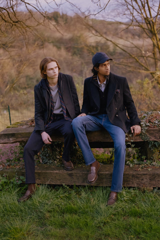 Two men sitting outdoors on a wooden bench wearing tailored jackets: one in a navy jacket with a striped scarf, grey t-shirt, and dark trousers with brown loafers; the other in a navy cap and double-breasted coat, blue jeans, and brown loafers