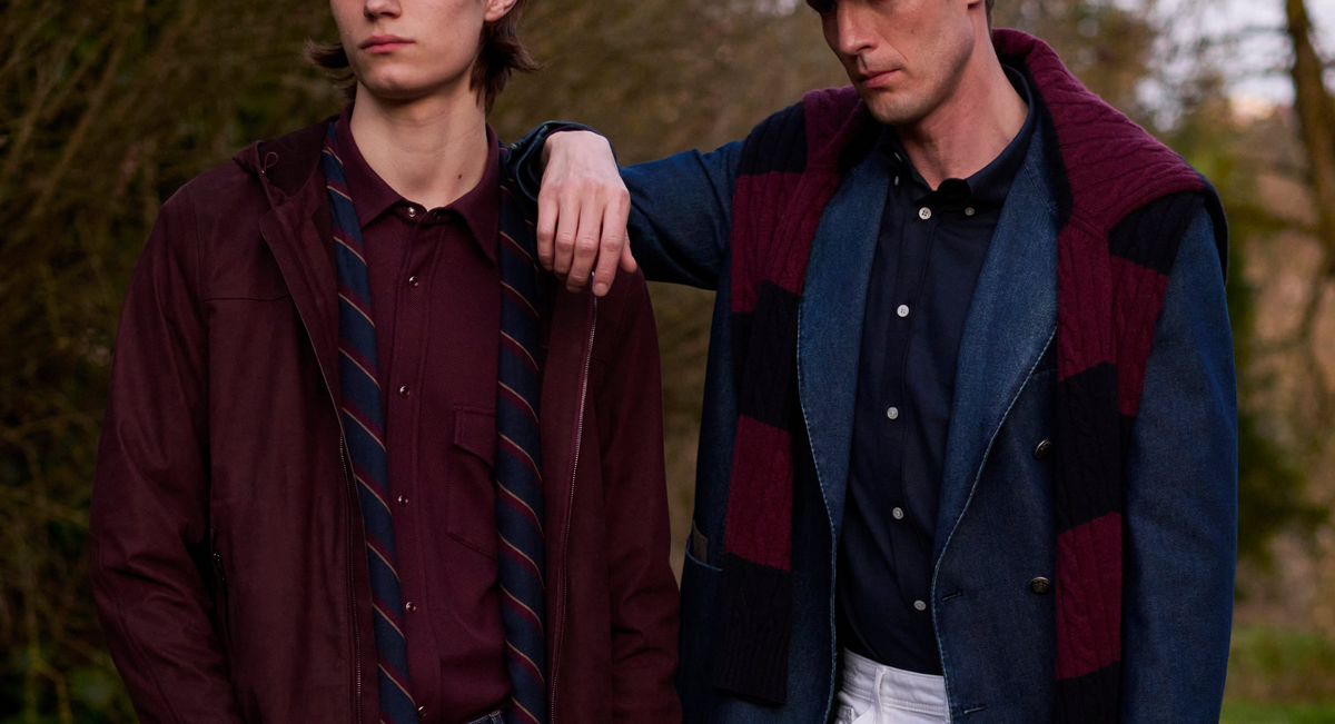 Two men outdoors wearing tailored menswear: one in a burgundy hooded jacket with a striped tie over a burgundy shirt and dark denim; the other in a denim blazer over a navy shirt and white trousers