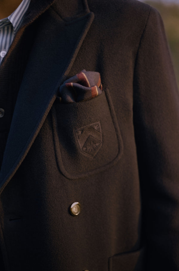 Close-up of a blue wool blazer with an embroidered crest patch pocket, patterned pocket square, and horn button, worn over a striped shirt