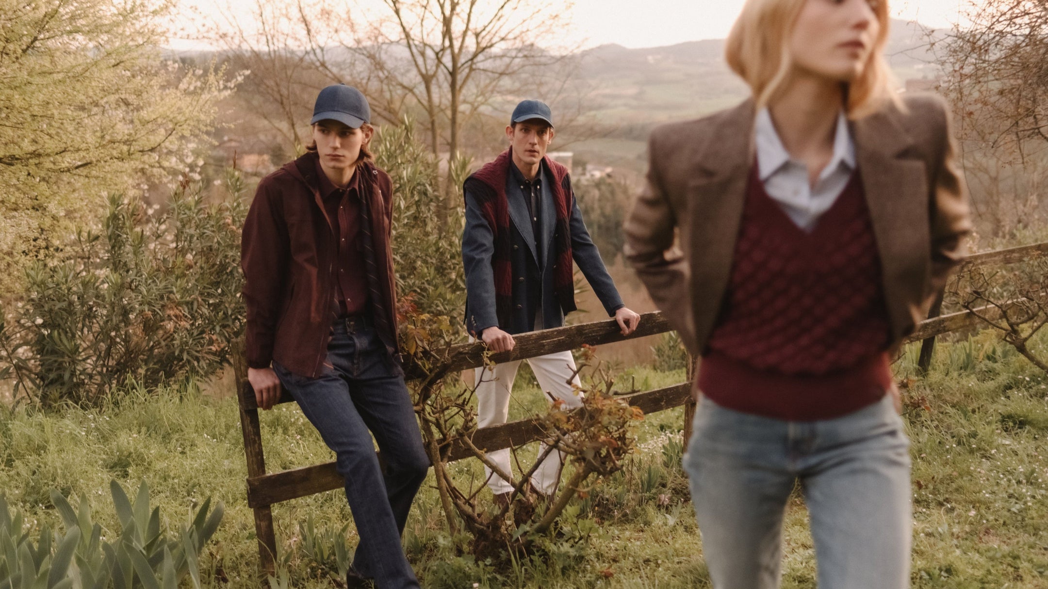 Three models in autumn-toned countryside setting: two men in flat caps, shirts and jackets lean against a wooden fence in denim and chino trousers, while a woman in a burgundy knit vest, blazer and jeans walks in the foreground