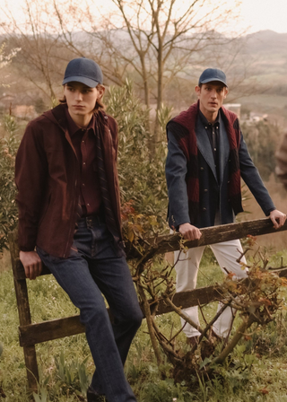 Two models in autumn-toned countryside setting: two men in flat caps, shirts and jackets lean against a wooden fence in denim and chino trousers
