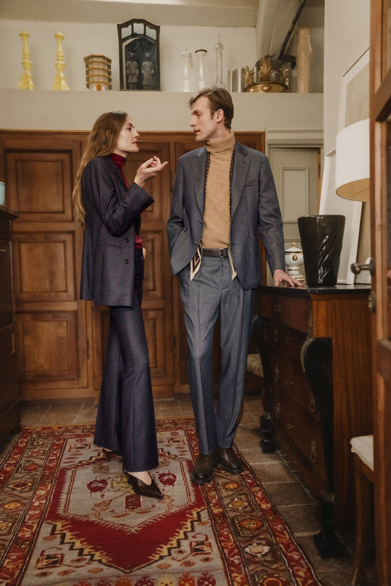 Two models in a rustic Italian interior with wooden paneling and a red patterned rug: a woman in a navy suit with flared trousers and a burgundy top, talking with a man in a grey-blue suit with a camel turtleneck and brown shoes.