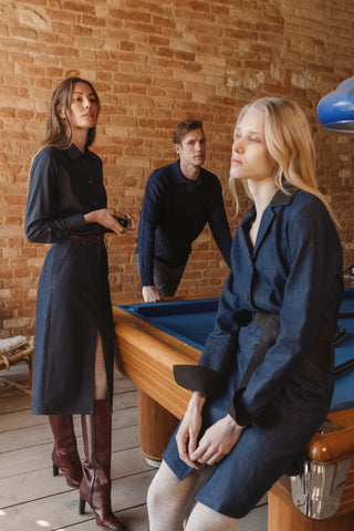 Two women and a man in tailored navy denim pieces gathered around a billiard table in a brick-walled room, one woman in a denim shirt and skirt with brown boots, the other in a navy denim coat, the man in a knit sweater leaning on the table