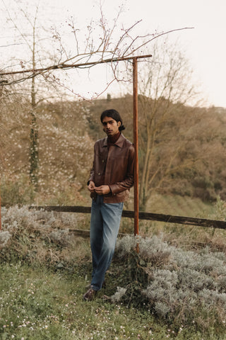 Man wearing a brown leather jacket over a turtleneck sweater, paired with light-wash denim jeans, standing outdoors in a countryside setting with blossoming trees and shrubs
