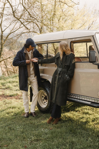 Man in a navy cap, dark navy coat, and cream trousers, and woman in a dark green belted trench leather coat with tan suede boots, standing beside a classic off-road vehicle on a grassy hillside