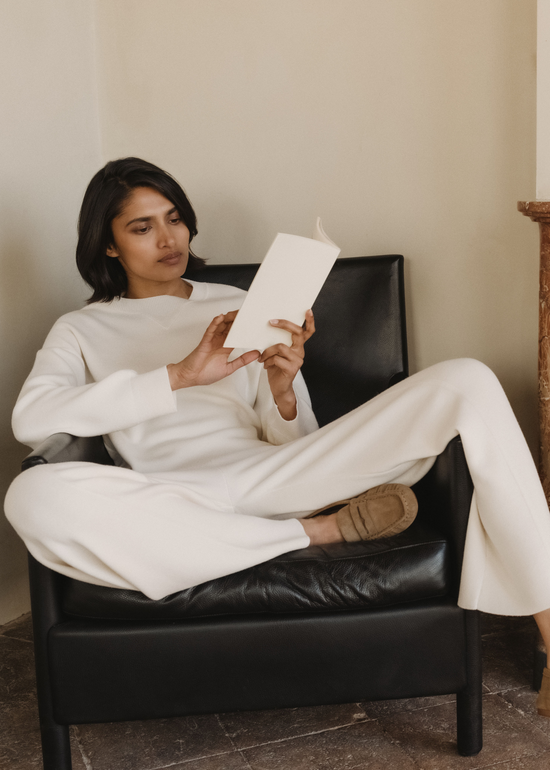 Female model in a cream knit sweater and wide-leg trousers with tan suede loafers, reclining in a black leather armchair and reading a book beside a marble fireplace in a sunlit room.