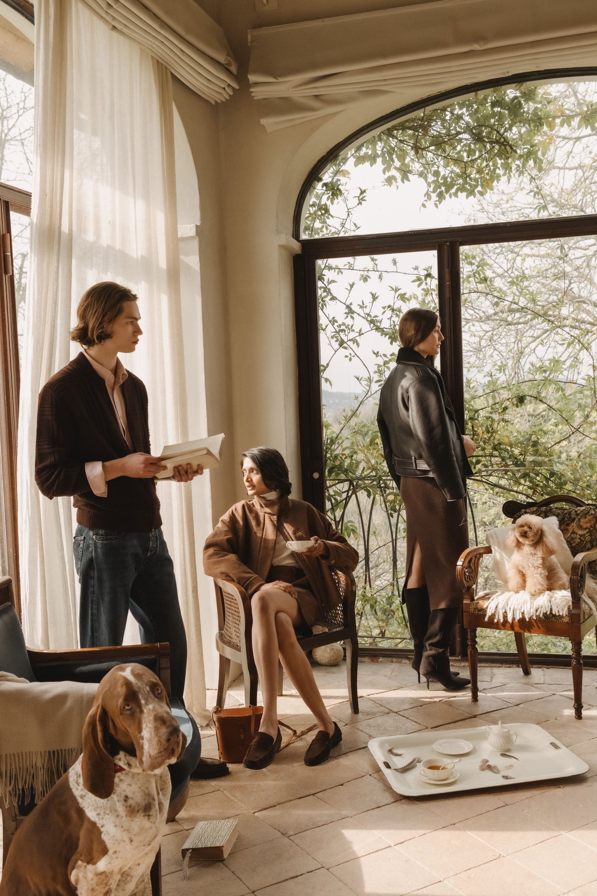 Three models in a sunlit room with an arched window: a man in a dark brown cardigan and jeans reading a book, a woman in a textured brown jacket seated with a teacup, and a woman in a brown leather jacket and brown midi dress standing by the window, with a basset hound and a small dog
