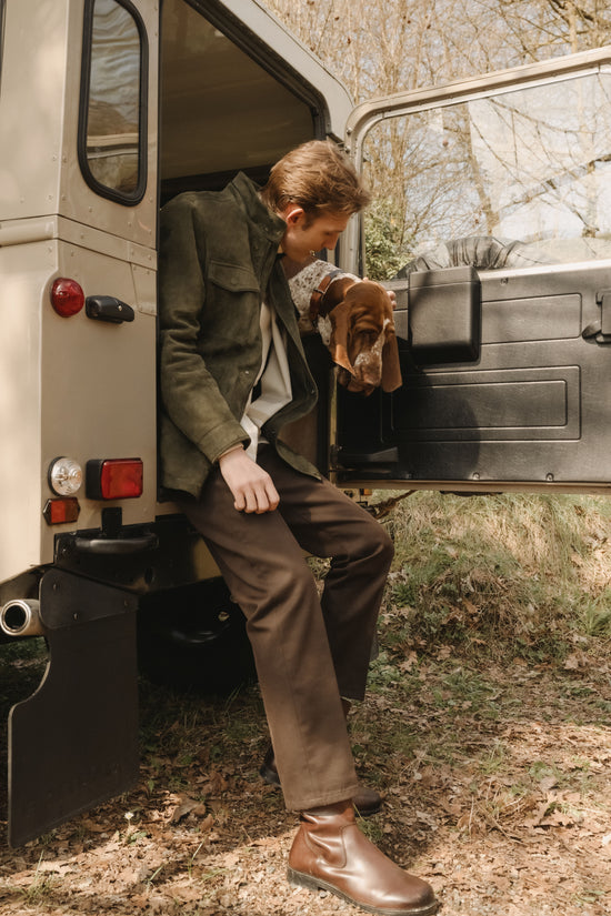 Male model in an olive green suede jacket, cream knit and brown trousers, paired with brown leather boots, stepping out of the back of a vintage off-road vehicle while a basset hound looks out from inside