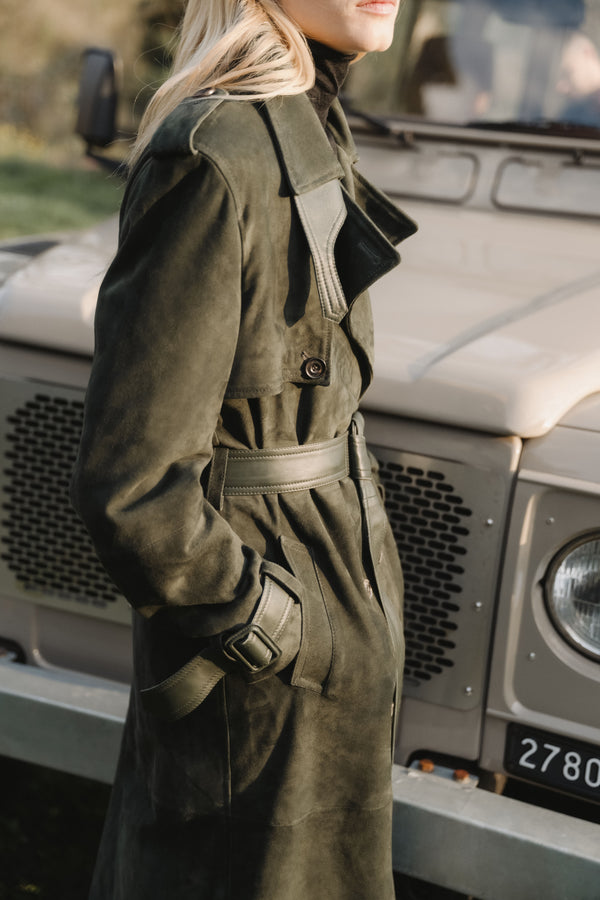 Woman wearing an olive-green suede belted trench coat with leather-trimmed collar, storm flap and cuff straps, styled with a dark roll-neck and standing in front of a classic off-road vehicle